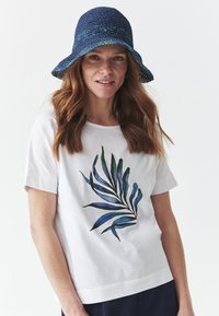 Chemise blanche à manches courtes avec un motif de feuilles bleues et vertes sur le devant, assortie à un chapeau seau tissé bleu marine avec une touche de bleu-vert.
