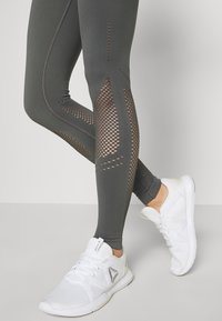 Ciemnoszare legginsy z siateczkowymi panelami wzdłuż boków, charakteryzujące się dopasowanym krojem i kontrastowym przeszyciem. W połączeniu z białymi butami sportowymi.
