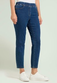 Pantalones vaqueros de mezclilla con un diseño ajustado, color azul oscuro, cintura elástica y dos bolsillos laterales. Combinados con zapatillas blancas para un look casual.