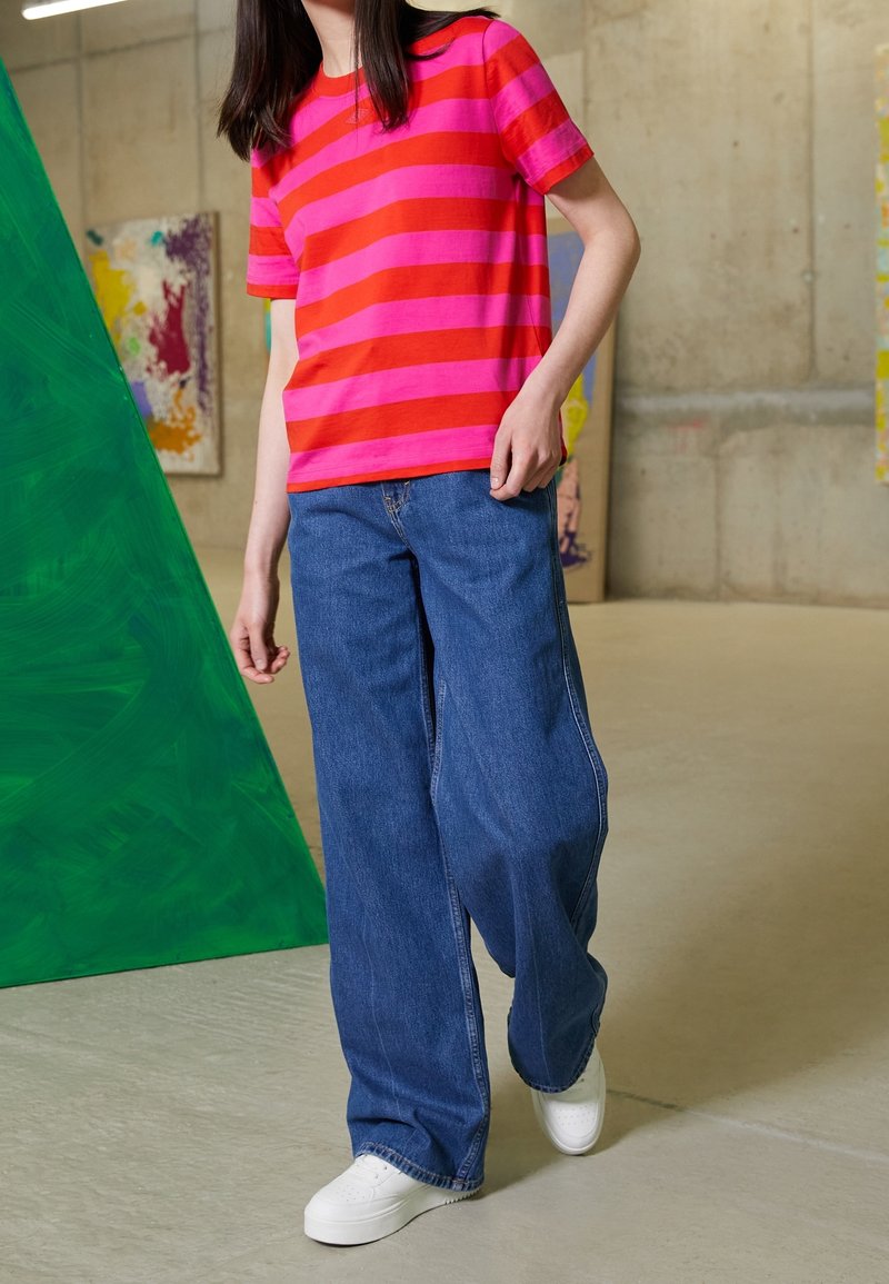 Personne portant une chemise rayée rouge et rose, un jean bleu large et des baskets blanches, debout près d'une sculpture triangulaire verte à l'intérieur.
