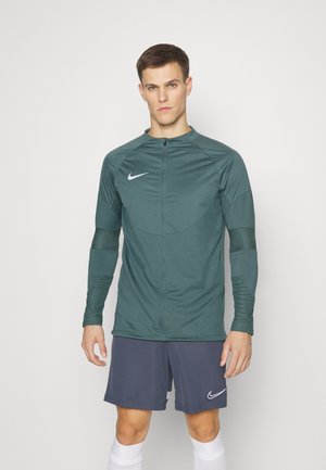 Donkergroene, lange mouwen, zip-up jack van lichtgewicht materiaal; heeft het Nike-logo en textuuraccenten op de mouwen. Gecombineerd met marineblauwe shorts.