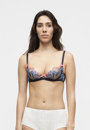 Woman with long dark hair wearing a sheer navy bra with pink and blue floral embroidery and white high-waisted underwear, facing forward.