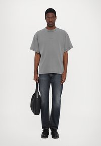 Grå oversized T-shirt med korta ärmar, matchad med mörkblåjeans. Modellen håller en svart läderväska och bär svarta skor.