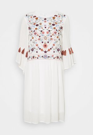 Robe blanche mi-longue avec des manches cloche trois-quarts, broderie florale sur le corps et franges brunes sur les manches.