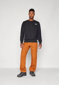 Zwarte sweatshirt met geribde manchetten, oranje broek met een gladde textuur en grijze schoenen. Logodetail op de sweatshirt.