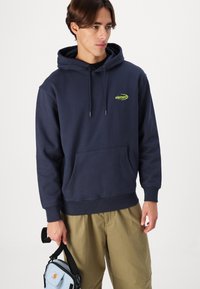 Navy blue hoodie with a front pocket, drawstring hood, and a bright green logo. Worn with light khaki pants, holding a small blue bag.