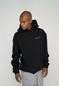 Black hoodie made of soft fabric, featuring a front kangaroo pocket and a small "MULTIPLY" logo in gray. Includes a drawstring hood.