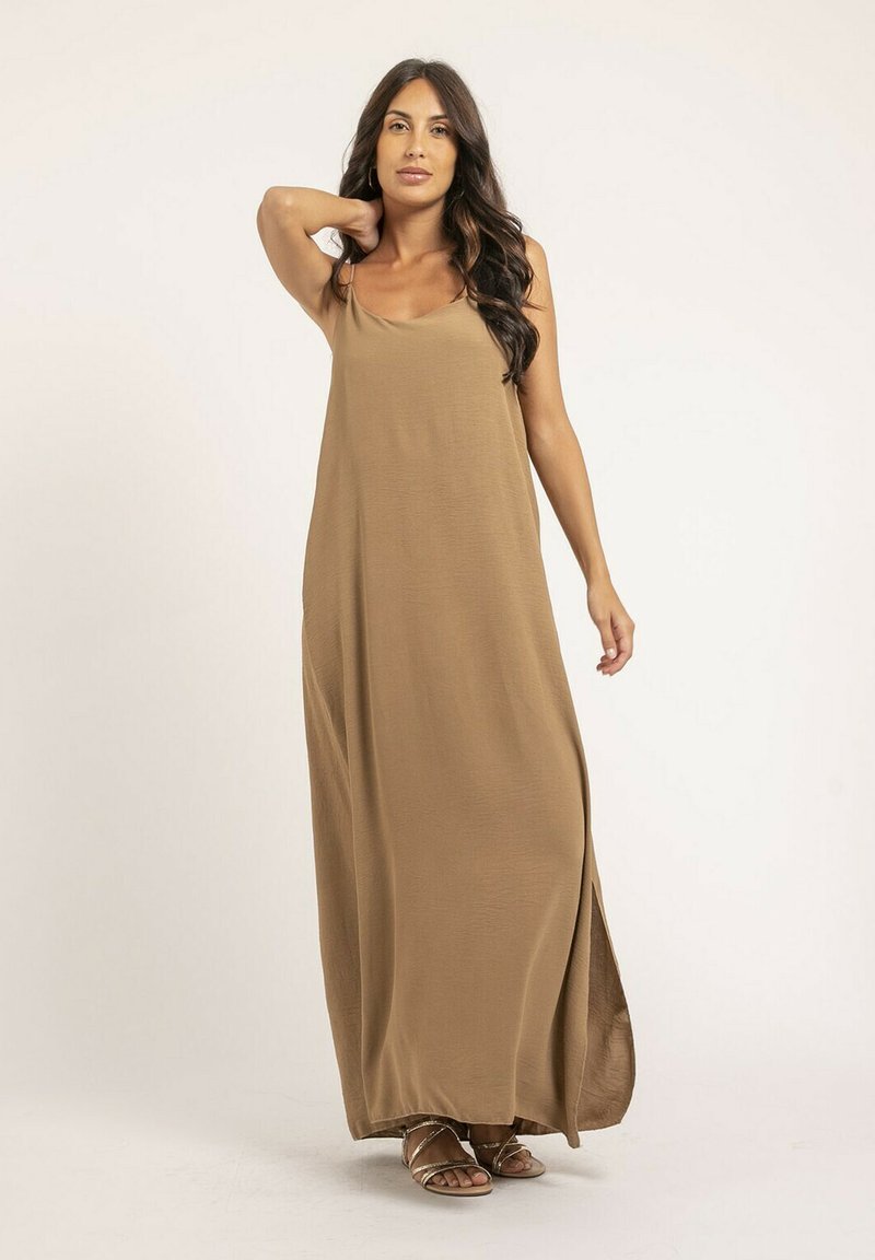 Robe maxi longue, sans manches, de couleur brune, fabriquée dans un tissu lisse, présentant une coupe décontractée et des fentes sur les côtés. Associée à des sandales à brides.