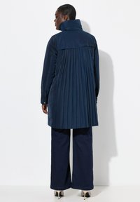 Cappotto trench navy con colletto alto e design con pieghe sul retro. Realizzato in materiale liscio, abbinato a jeans a gamba larga scuri e tacchi.