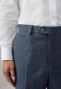 Dunkelblaue, strukturierte Hose mit einem dezenten Glitzern, ausgestattet mit einem Knopfverschluss, seitlichen Taschen und einem taillierten Schnitt. Ein weißes Hemd ist teilweise sichtbar.
