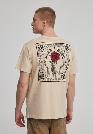 T-shirt in cotone beige con un grande design grafico nero che presenta una rosa rossa, mani ed elementi decorativi sulla parte posteriore.