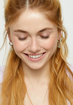 Joven con cabello largo y rojo y pendientes plateados sonríe suavemente con los ojos cerrados sobre un fondo claro y sencillo.
