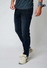 Tmavě modré džíny z denimu s úzkým střihem, jemnými švy a předními kapsami, doplněné světlými sportovními botami.