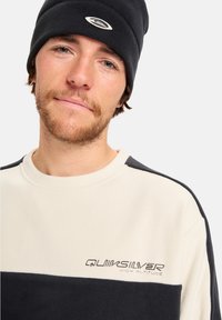 Vīrietis ar gaišu bārdu, tērpies melnā beanie cepurē un melnbaltajā Quiksilver vilnas kreklā ar logo uz krūtīm.