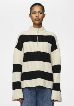 Pull en tricot avec un col haut, présentant des rayures horizontales alternées noires et crème, un tissu texturé et une fermeture éclair à l'avant.