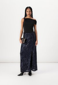 Melna off-plecu blūze, ko kombinē ar tumši zilas sekvīnu maksi svārki ar priekšējo šķēlumu, papildināta ar mazu melnu clutch somiņu un melniem apaviem ar augstiem papēžiem.