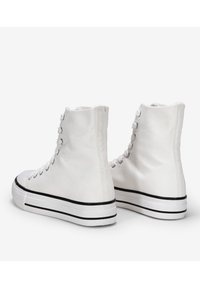 Zapatillas altas de lona en blanco, con una suela de goma negra que tiene dos rayas y ojales de metal para los cordones.