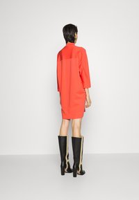 Korallenfarbenes Kleid mit Dreiviertelärmeln, lockerer Passform und kontrastierendem Rückenteil, kombiniert mit schwarzen kniehohen Stiefeln mit goldenen Akzenten.