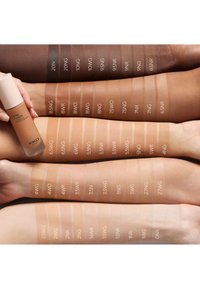Vier Arme mit Foundation-Proben in verschiedenen Farbtönen, die mit Codes beschriftet sind, gehalten neben einer KIKO Milano Love Fusion Foundation-Flasche.
