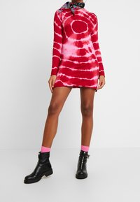 Vestido mini de manga larga en tie-dye rojo y rosa con una silueta ajustada. Combinado con botas negras hasta el tobillo y calcetines rosas. Textura suave.