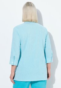 Camicetta a righe blu chiaro con maniche a tre quarti, dettagli con bottoni e un design morbido. Il tessuto appare liscio e con una texture.