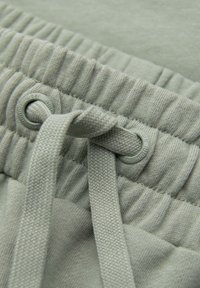 Gros plan d’une ceinture élastique gris clair avec un cordon texturé passé à travers des œillets métalliques sur un pantalon en tissu doux.