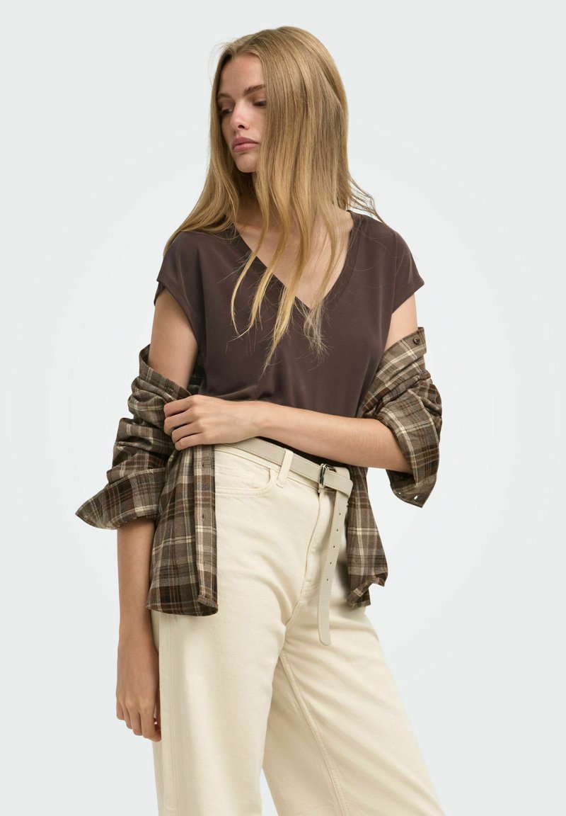 Femme blonde portant un haut marron, un pantalon beige avec ceinture et une chemise à carreaux drapée sur ses épaules, regardant sur le côté.
