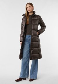 Langer Puffer-Mantel in Dunkelbraun, mit hohem Kragen und vertikalem Steppmuster. Kombiniert mit weiten Jeans und dunklen braunen Stiefeln.