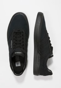 Svarta canvas sneakers med rund tå, svart gummisål och snörning. Har tre horisontella ränder på sidan och logotyp på tungan.