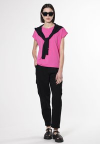 Roze T-shirt met korte mouwen gecombineerd met zwarte cargobroek. Een zwarte trui gedrapeerd over de schouders. Zwarte chunky sandalen maken de outfit compleet.