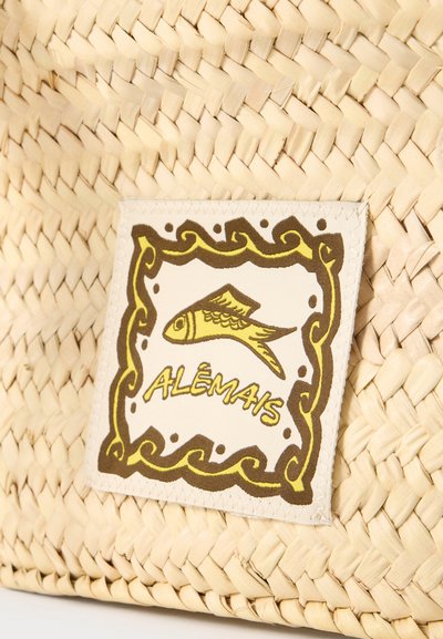 Texture de paille tissée avec une étiquette en tissu cousue montrant une illustration de poisson jaune et le mot « ALÊMAIS » entouré d’une bordure décorative marron et jaune.