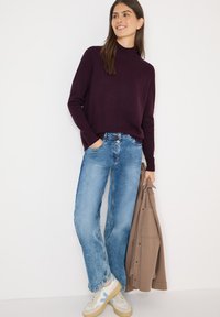 Dunkellila Strickpullover, hellblaue Jeans mit weitem Bein, beige Sneakers mit blauen Akzenten. Braune Jacke in einer Hand gehalten. Einfarbiger weißer Hintergrund.