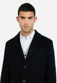 Jonge man met kort donker haar, gekleed in een donkerblauwe jas over een lichtblauw gestreept shirt en een grijze cardigan, tegen een effen achtergrond.