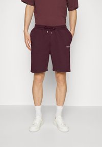 Homme portant un short bordeaux foncé avec cordon de serrage, une chemise assortie, des chaussettes blanches et des baskets blanches, debout devant un fond blanc.