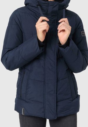 Granatowa pikowana kurtka z wysokim kołnierzem i sznurkami, noszona przez osobę regulującą kaptur, wyposażona w zapinane na zamek boczne kieszenie i naszywkę z logo na rękawie.