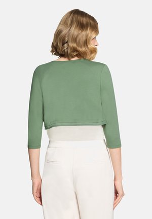 Frau mit schulterlangem, welligem blondem Haar trägt ein bauchfreies grünes Oberteil mit Dreiviertelärmeln und hoch taillierte weiße Hosen, von hinten gesehen.