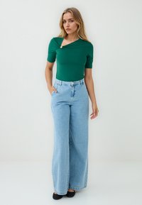 Vrouw met lang, golvend haar poseert in een groene top en losse blauwe jeans, met handen in de zakken, tegen een lichte achtergrond.