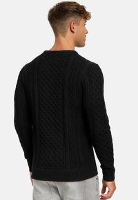 Schwarzer Kabelstrickpullover mit strukturiertem Muster, rundem Halsausschnitt und gerippten Bündchen und Saum. Kombiniert mit grauen Hosen.