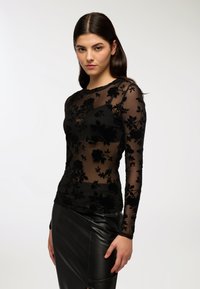 Top nero a maniche lunghe realizzato in tessuto trasparente con applicazioni in velluto floreale, abbinato a una gonna a tubino in pelle nera. Texture liscia, design aderente.