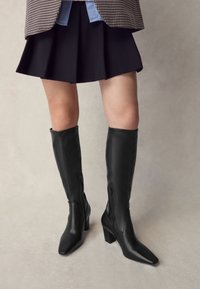 Svarta knähöga stövlar med klackar i slätt läder, med spetsig tå och sidodragkedjor, kombinerade med en kort svart kjol.