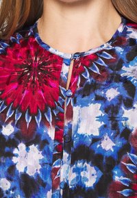 Blouse colorée à manches longues avec un motif tie-dye rouge, bleu et noir. Elle a un col rond et des boutons décoratifs.