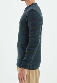 Pull rayé bleu foncé avec poignets et ourlet côtelés. Le tissu semble texturé avec des nuances variables. Le modèle porte un pantalon beige clair.