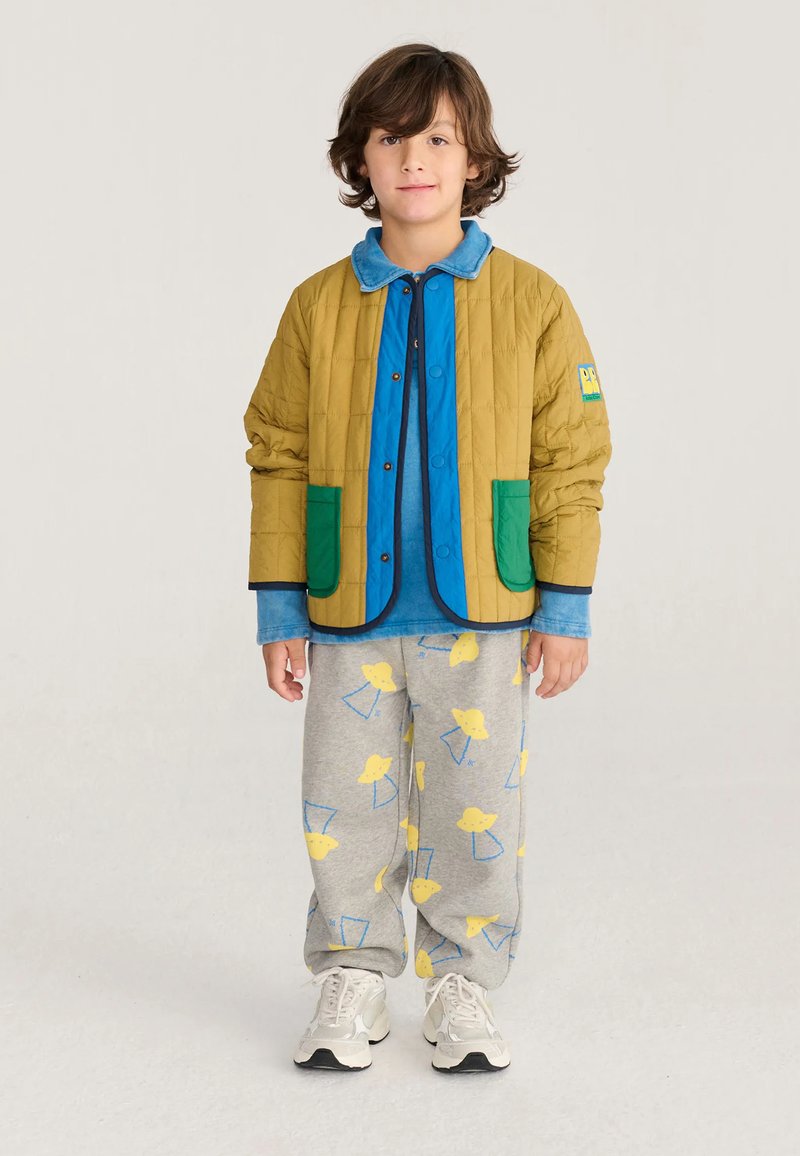 Enfant aux cheveux bruns mi-longs portant une veste moutarde avec des poches vertes, une chemise bleue, un pantalon gris à motifs et des baskets blanches.