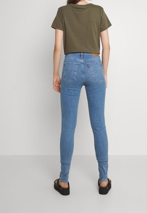 Femme portant un t-shirt vert olive, un jean skinny taille haute bleu clair et des sandales à plateformes noires, debout de dos face à la caméra.