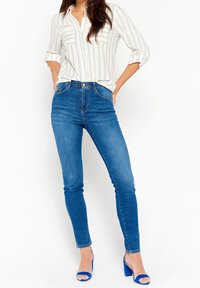 Modré džínové skinny jeans s vysokým pasem, doplněné bílou pruhovanou košili s dlouhým rukávem. Boty jsou modré sandály na podpatku.