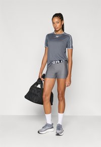 Atlétikai outfit szürke rövid ujjú pólóval, szürke rövidnadrággal mintás derékpánttal, valamint szürke tornacipőkkel. A modell egy fekete edzőtáskát tart a kezében.