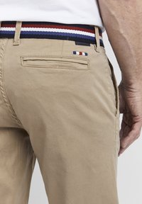 Bež pamukove chinos hlače s stražnjim džepom i tkanim remenom u tamnoplavoj, crvenoj i bijeloj prugastoj kombinaciji. Glatka tekstura, ležeran dizajn i minimalni detalji.