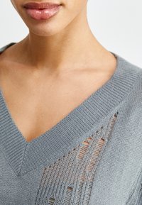 Grå stickad tröja med V-ringning, med ett strukturerat mönster och subtila utskärningar längs halsringningen. Mjuk, lätt tyg.