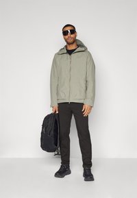 Gray zippered jacket with a hood, textured fabric, worn over a black top and black pants. Black backpack in hand, black athletic shoes.
