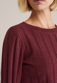 Burgundy neule, jossa on ribbitekstuuri ja pystysuorat raidat. Ominaisuuksiin kuuluu pyöreä pääntie ja pitkät hihat. Pehmeää neulosta.